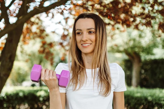 Una donna sorridente si allena con un manubrio viola in un parco luminoso